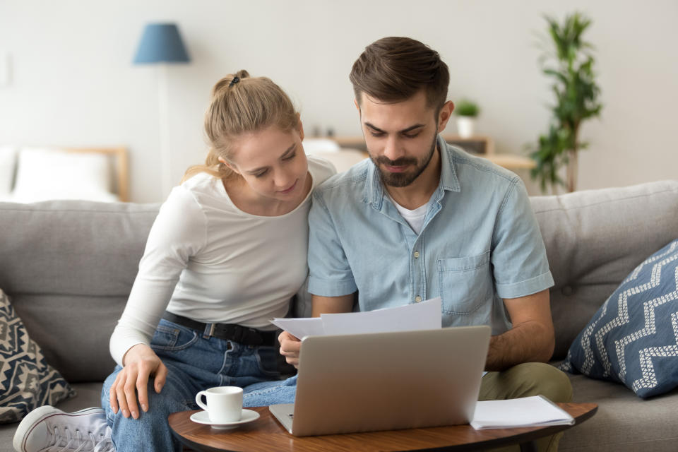 Young serious couple holding reading financial documents checking paying household bills online on laptop, managing finances taxes, reviewing bank accounts, planning budget expenses at home together