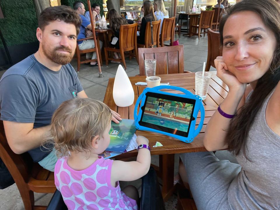 A family taking a selfie while at a restaurant table with a child watching an ipad.