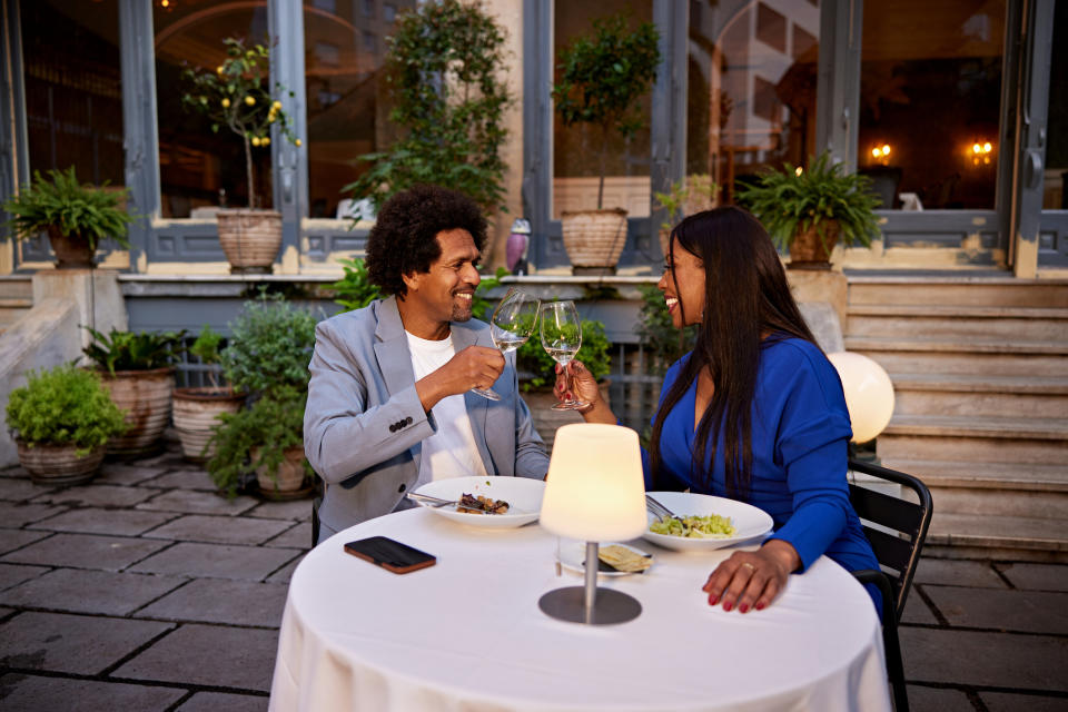 A couple is dining at an outdoor restaurant, toasting with glasses of wine throughout their meal. They are smiling and seem to be enjoying each other's company.