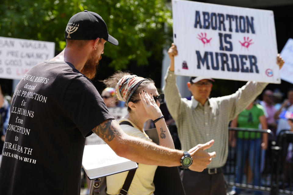 Abortion opponent Alexander Wirth, left, reads from the Bible as he's confronted by a woman supporting abortion rights, center, outside the South Carolina Statehouse on Thursday, July 7, 2022, in Columbia, S.C. Protesters clashed outside a legislative building, where lawmakers were taking testimony as they consider new restrictions on abortion in the wake of the U.S. Supreme Court's decision overturning of Roe v. Wade. (AP Photo/Meg Kinnard)