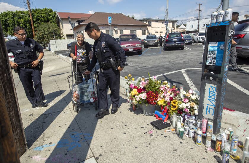 LINCOLN HEIGHTS, CA -SEPTEMBER 12, 2022: LAPD officers Nicholas Torres, left, and Erick Rodriguez help a blind man (didn't want to give his name) who lives in the neighborhood, make his way past a memorial for Winfield Mikey Lee, 17, on Workman St. in Lincoln Heights, one two teenage boys who were shot and killed Sunday night at a street carnival in Lincoln Heights.The LAPD officers were keeping an eye on activity around the memorial. (Mel Melcon / Los Angeles Times)