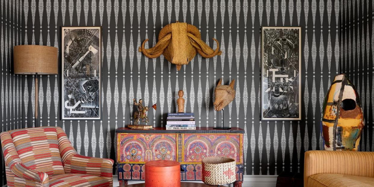 a sitting room has dark gray and white patterned wallpaper, a sofa, upholstered chair, cocktail table, moroccan console, a standing ceramic sculpture, and on wall a palm leaf water buffalo head and works on paper