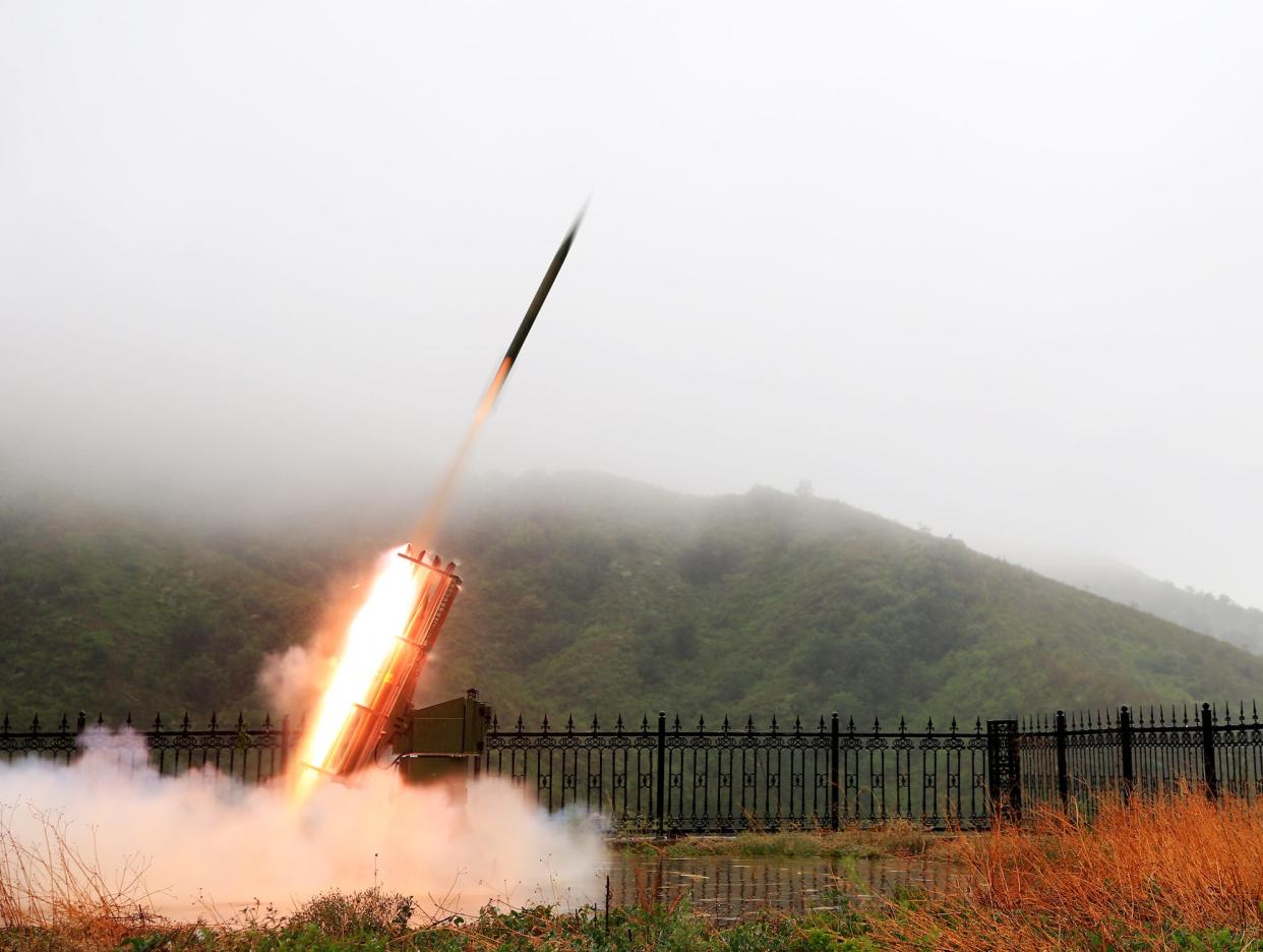 thin long rocket launches toward the sky with fiery flare from a cage-like device on the ground with green hills in the background