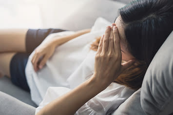 A woman sits on a couch while holding her head. 