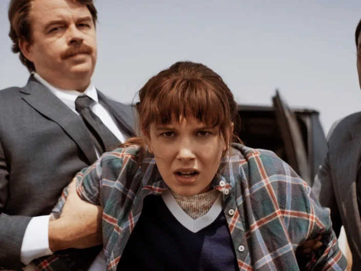millie bobby brown as eleven in stranger things, leaning forward while two men in suits hold onto her arms, pulling her back into a car