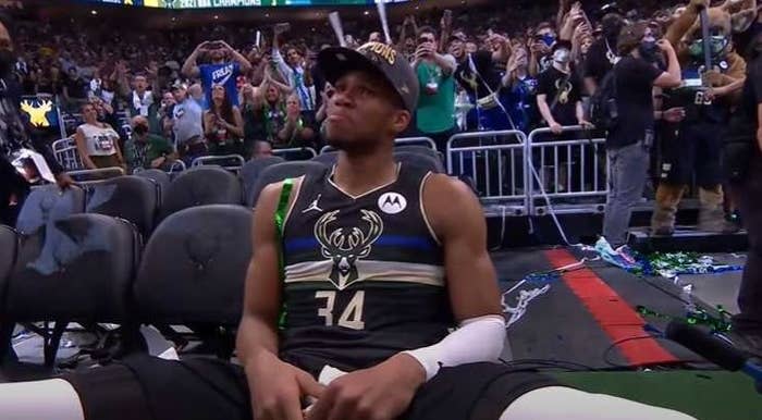 Emotional Giannis Antetokounmpo sits in a chair while wearing a champions hat