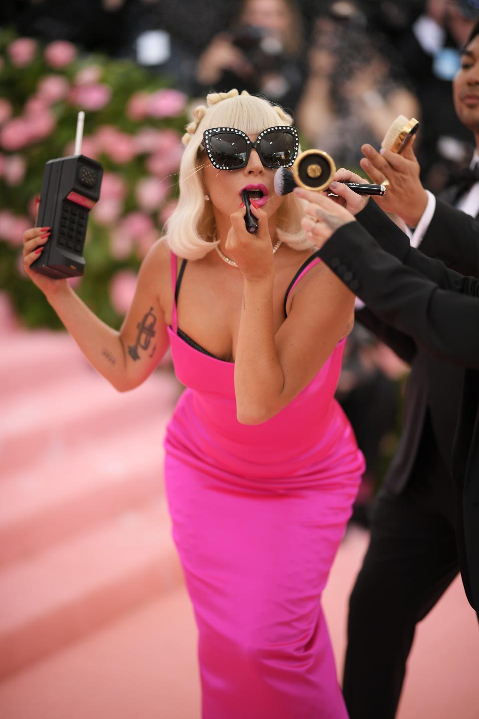 Lady Gaga, wearing a tight hot pink dress, applies lipstick and holds a large phone prop while men in suits hold hair and makeup brushes to her head.