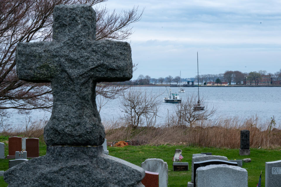 A stone cross and gravestones on City Island looking over to Hart Island, with small tugboats on the water.