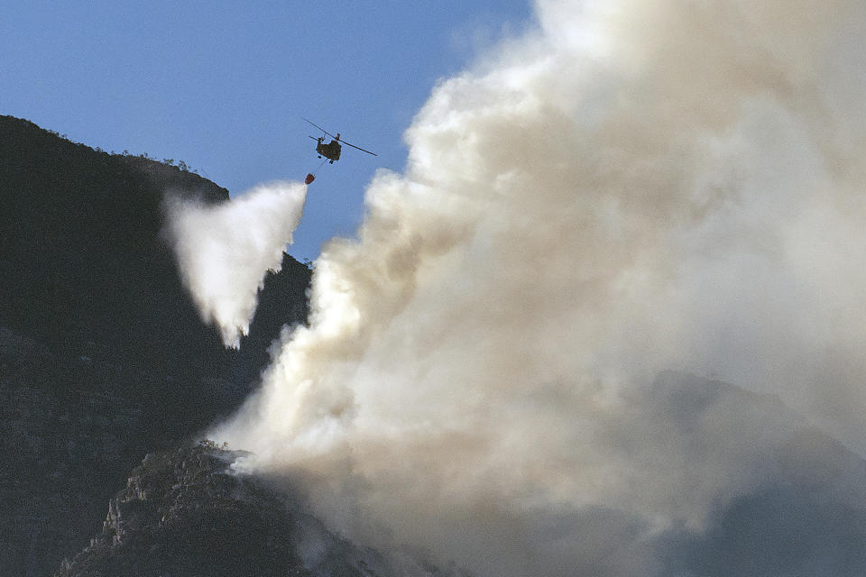 A South African military helicopter drops water on the top of Table Mountain in Cape Town, South Africa, Tuesday, April 20, 2021. A massive fire spreading on the slopes of the city's famed Table Mountain is kept under control as firemen and helicopters take advantage of the low winds to contain the blaze. (AP Photo/Jerome Delay)