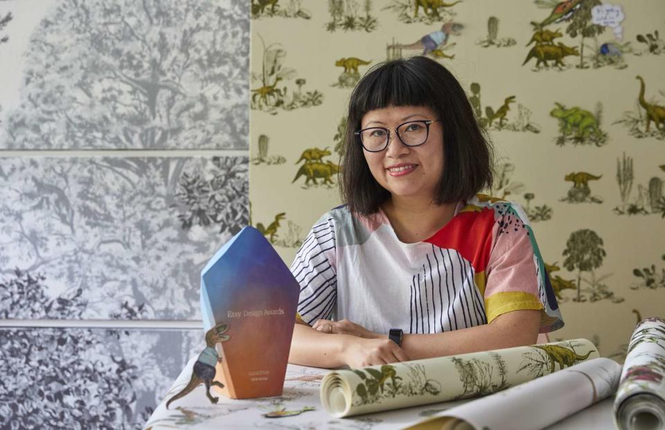 Sian Zeng sits in front of a wall decorated with dinosaur print green wallpaper with a pink and purple award on her desk