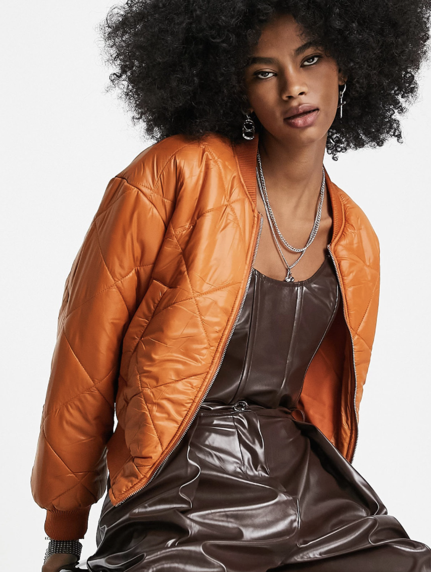 Woman with dark curly hair wears an orange Bershka quilted bomber jacket over a brown dress.