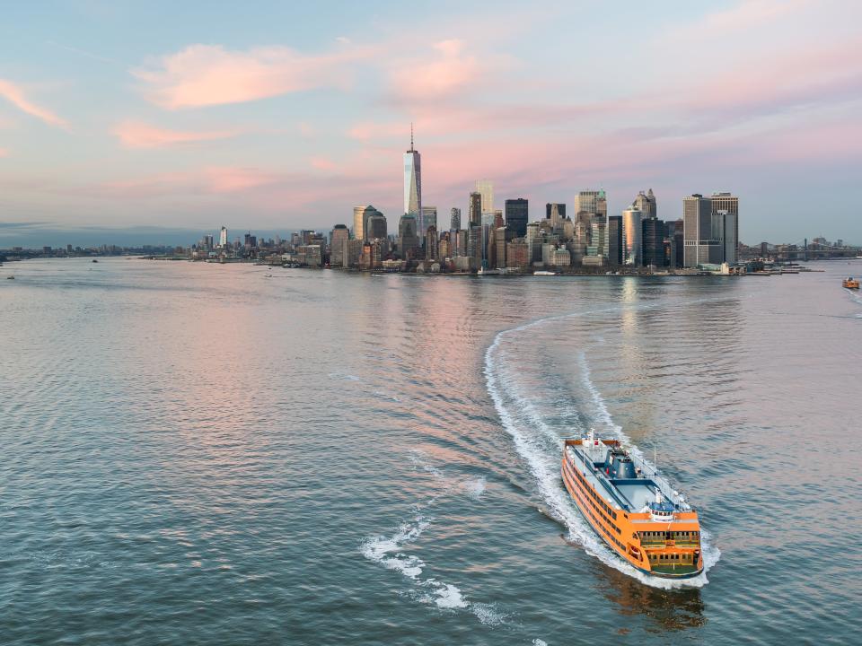 This aerial view from a helicopter shows the southern tip of Manhattan at sunset. The Hudson River is on the left and the East River is on the right. Seen here also is One World Trade Center, the tallest building in the western hemisphere. Also seen here is the orange colored Staten Island Ferry.