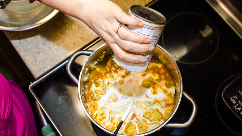 A hand pouring a can of coconut milk into a pot of soup