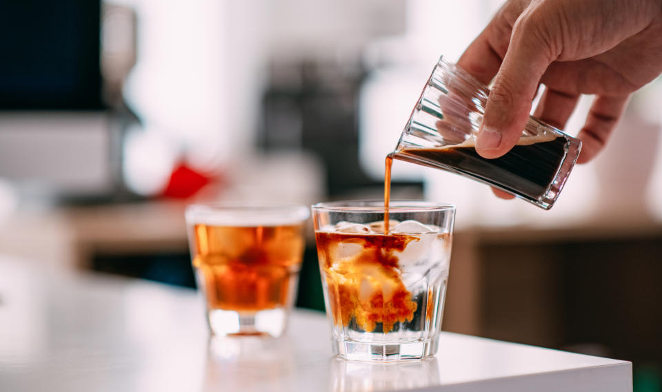 Hand pouring coffee from small pitcher into glass with ice; another iced coffee in background