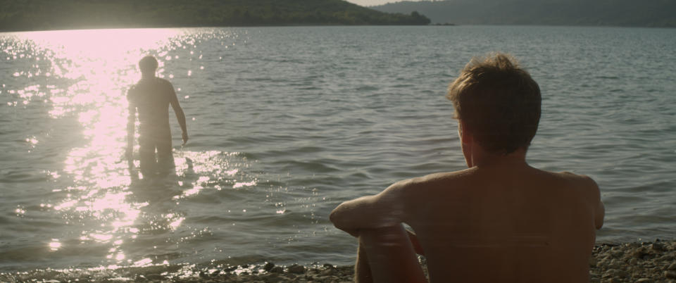 Two people at a lakeside: one sits on the rocky shore, and the other walks into the water, backlit by the sun. Their names are unknown