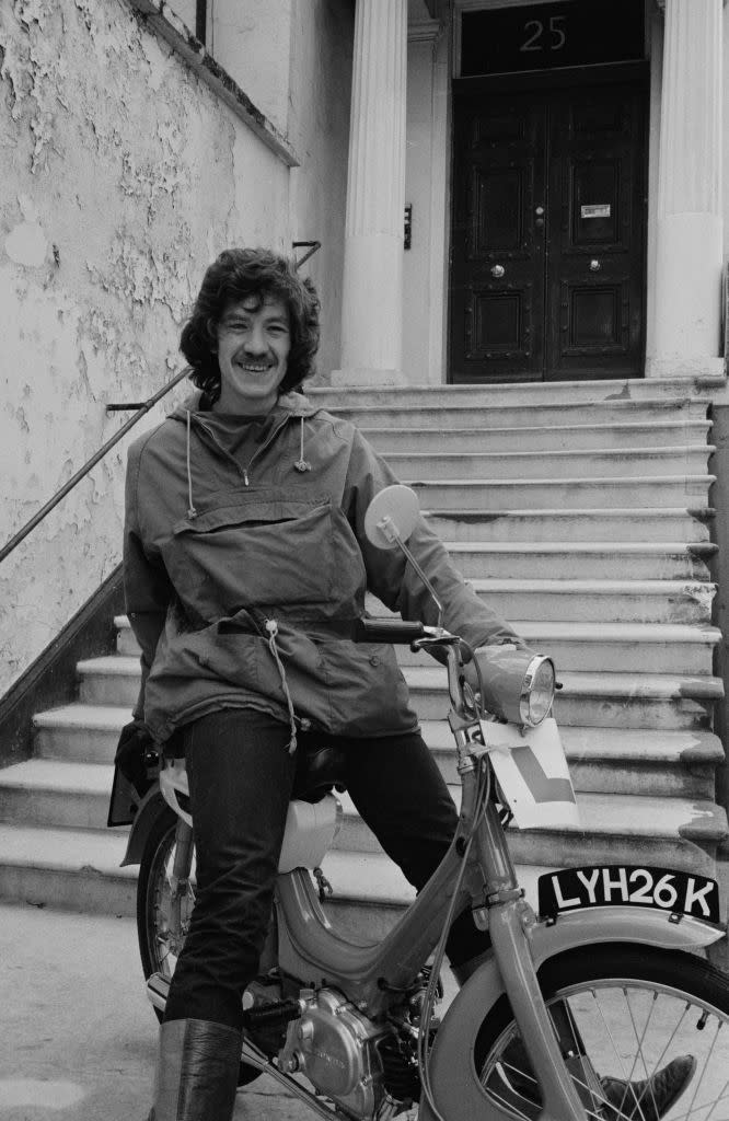 Person is sitting on a motorcycle, smiling, wearing a casual jacket and pants, parked in front of steps leading to a door