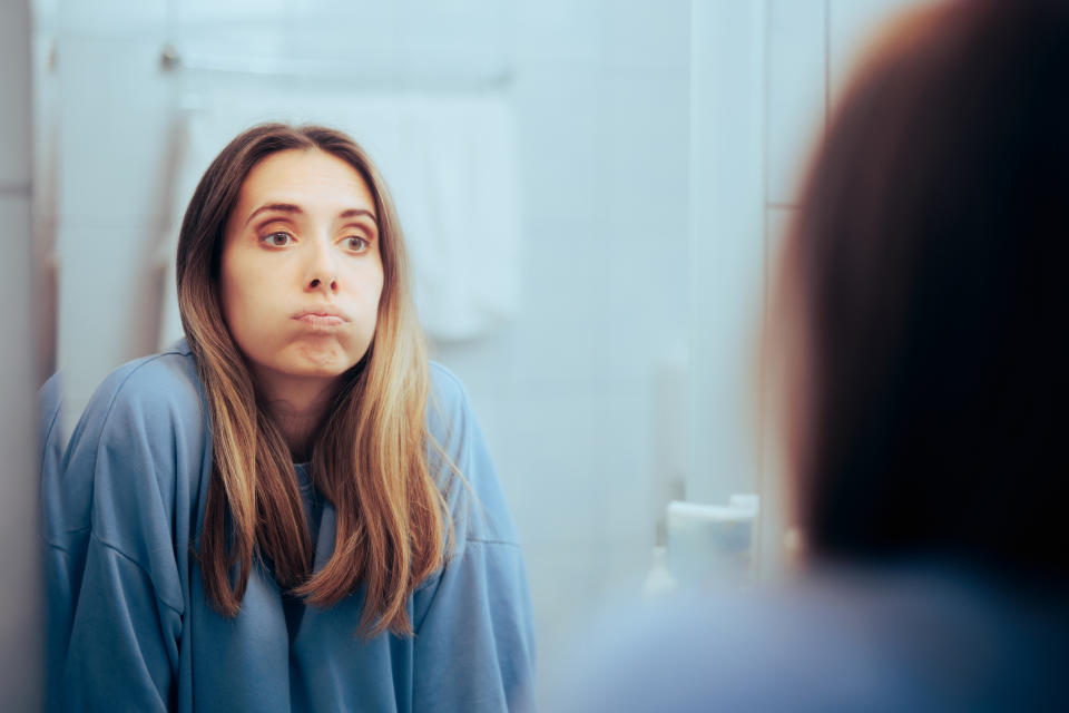 Woman in a cozy sweatshirt looking at herself in the bathroom mirror, puffing her cheeks with a thoughtful expression. No text in the image