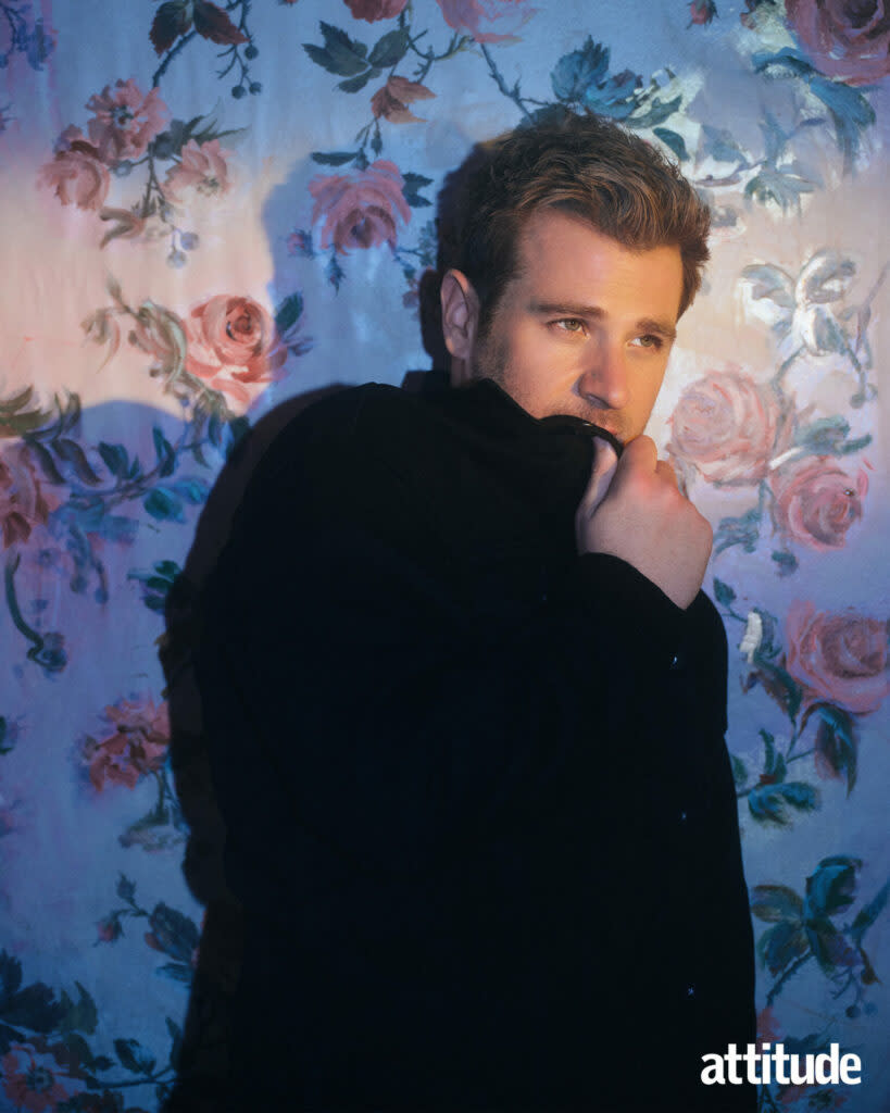 Scott is standing against floral wallpaper with a black jacket covering his mouth.