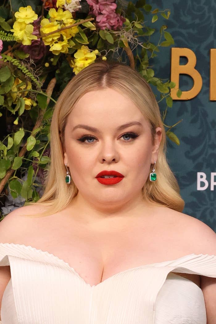 Nicola Coughlan wearing an off-the-shoulder dress with statement earrings stands in front of a floral backdrop at a public event