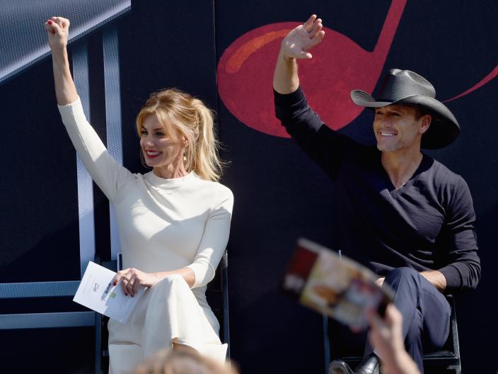 Faith Hill in a white dress and Tim McGraw in a black suit and cowboy hat onstage waving to their right