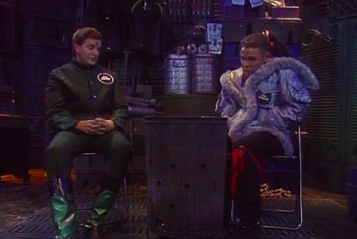 Rimmer and Lister sit in front of a fire in a barrel in a spaceship
