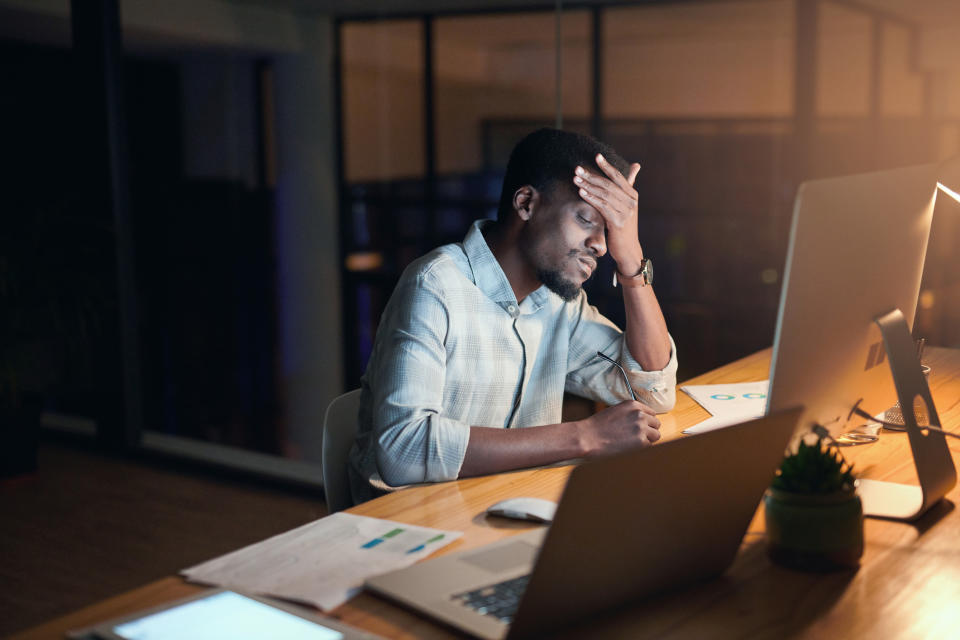 A man in a dress shirt sits at a desk, holding his head in his hand, appearing stressed and exhausted while looking at computer screens and documents