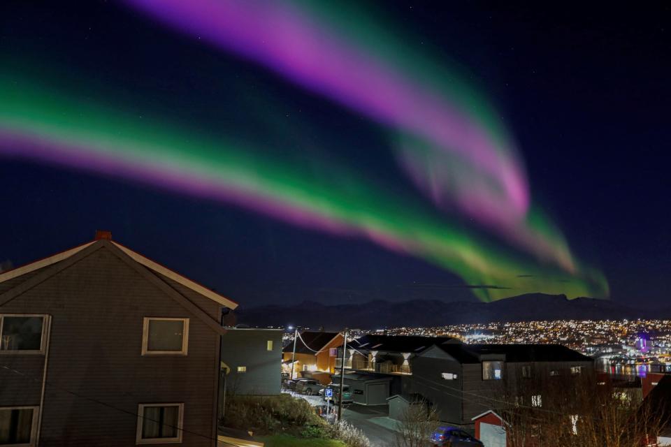 ribbons of green-pink-purple northern lights stretch across the night sky above houses and lights of a vast city