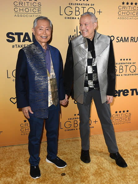 George Takei and Brad Altman holding hands on the red carpet at the Critics Choice Association Celebration of LGBTQ+. Both are wearing stylish vests