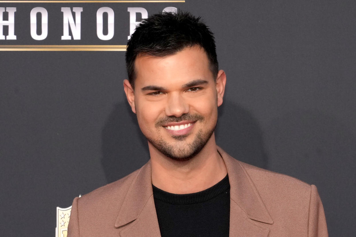  A headshot of Taylor Lautner wearing a brown suit jacket looking at the camera and smiling. 