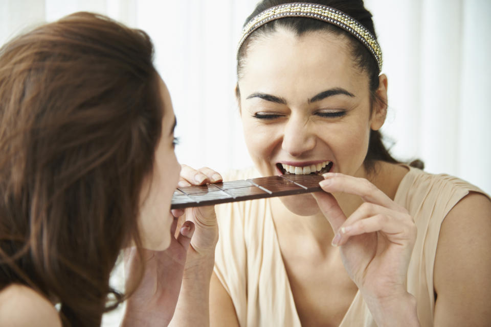 Two women share a chocolate bar, smiling and biting it from opposite ends. Both have dark hair; one wears a headband