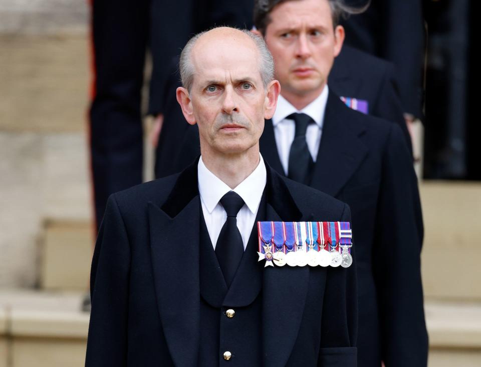 a man stands at the front of a line with a neutral expression on his face, he wears dark navy suit and several medals