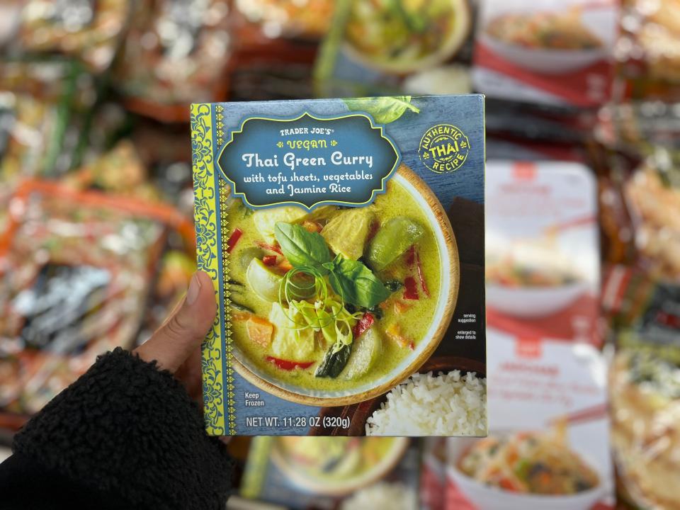 A hand is holding a green and blue box with a photo of Thai green curry with tofu, vegetables and rice on it