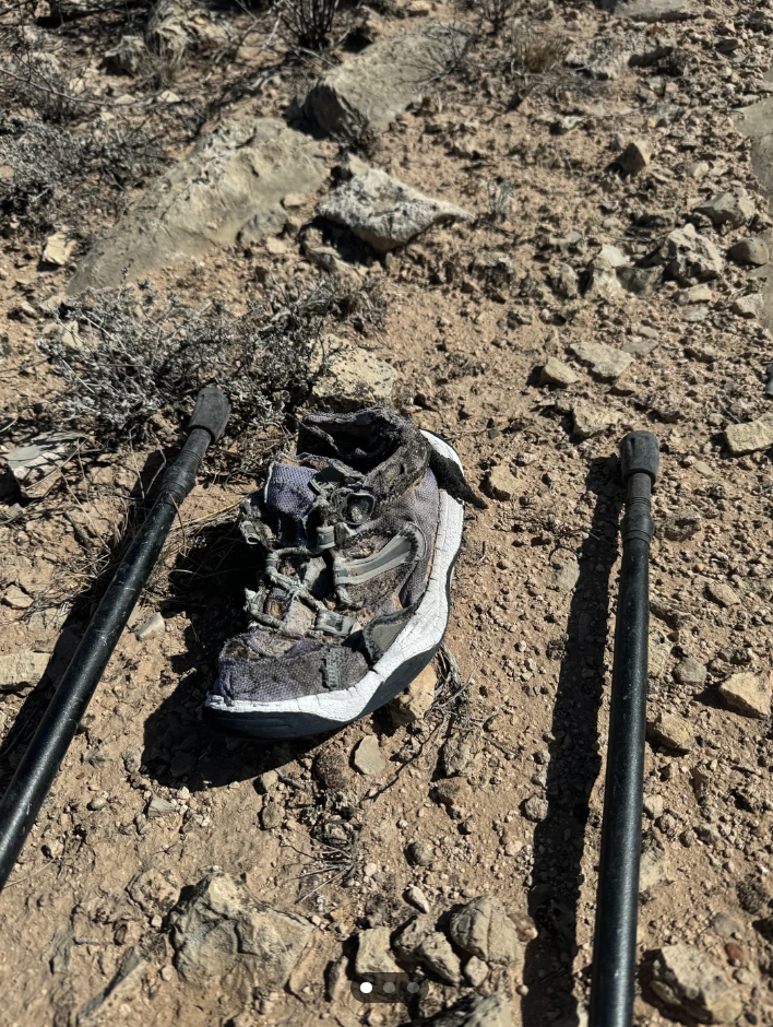 Worn-out shoe on rocky ground between two trekking poles, suggesting a hiker's challenging journey