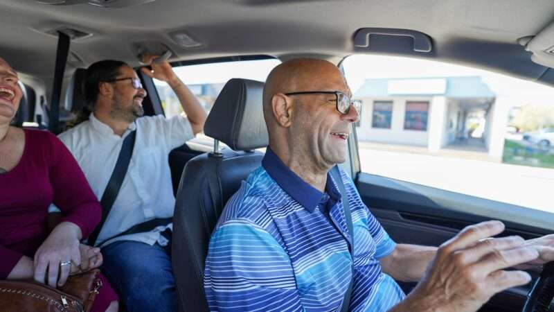 Inside a minivan, a man in a blue polo shirt drives two passengers in the back seat, and all three are smiling.