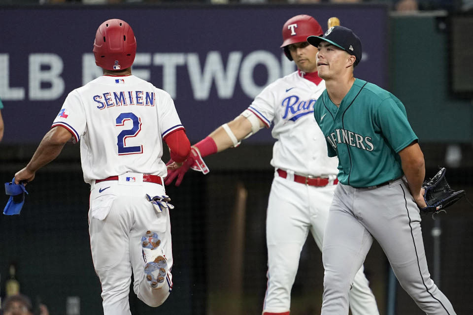 Seattle Mariners starting pitcher Bryan Woo, right, backs up the plate as Texas Rangers' Marcus Semien (2) celebrates with Nathaniel Lowe, center, after scoring on a Corey Seager single in the first inning of a baseball game, Saturday, June 3, 2023, in Arlington, Texas. (AP Photo/Tony Gutierrez)