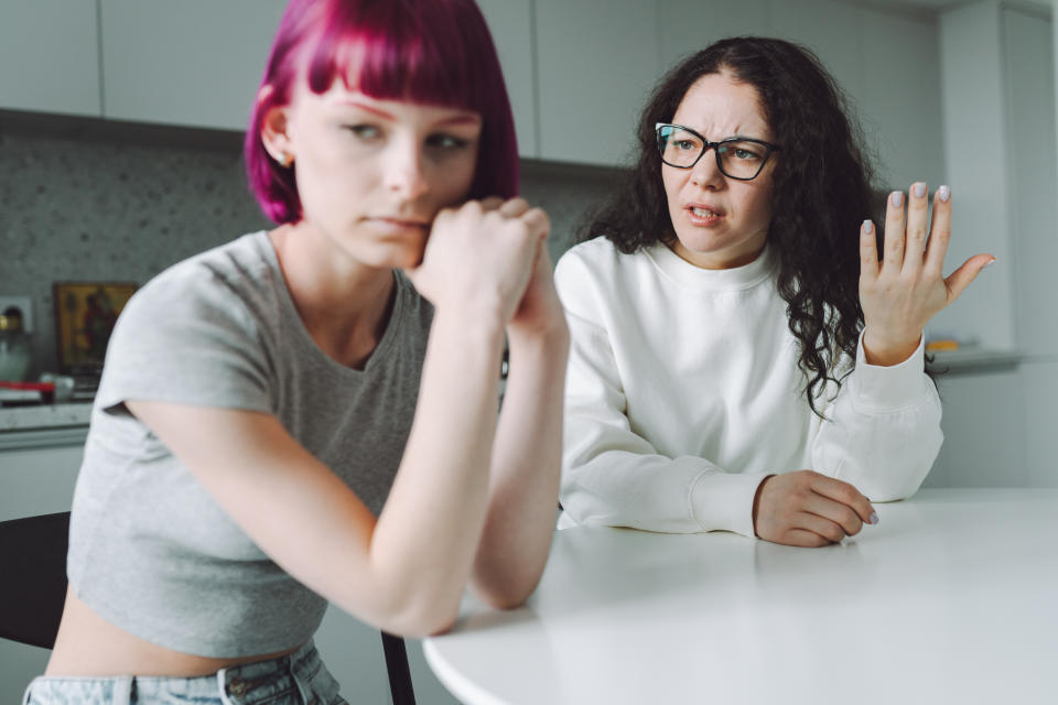 Teen with pink hair in a grey crop top sitting at a table, looking away, while a woman with glasses and curly hair in a white shirt gestures frustratedly