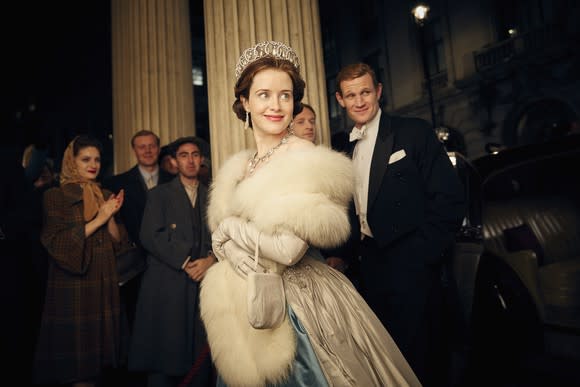 A scene from Netflix original "The Crown" with Claire Foy and Matt Smith, showing a striking young woman smiling, wearing a stole and a crown, with a man in a tuxedo in the background.