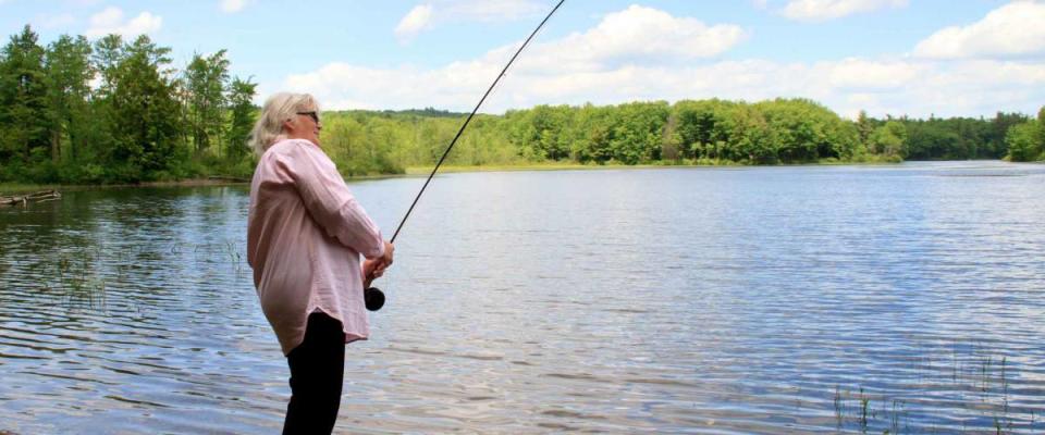 Elderly, senior citizen fly fish and catching fish in mountain wilderness lake. Woman is fighting a fish, blue sky clouds, forest background, wind and small waves on the water.