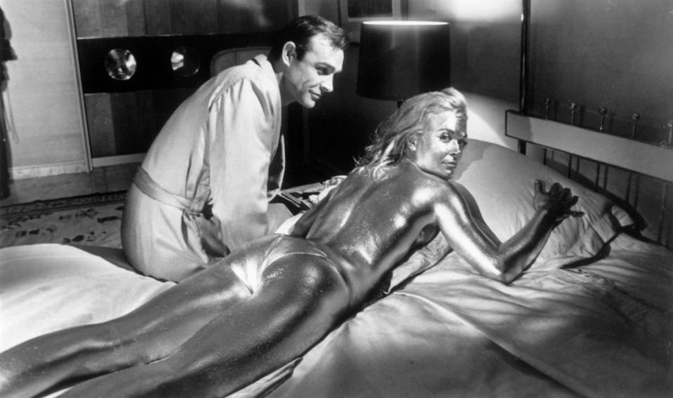 <p>Sean Connery sitting beside his co-star English actress, Shirley Eaton, covered in gold during the filming of a scene from <em>Goldfinger.</em></p>