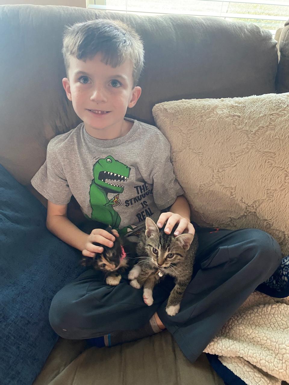 Bradley Lewis, age 7, sits on his couch with sibling cats Lily (pink collar) and Snowball in their home in Katy, Texas. The family used a 2021 Ford F-150 Hybrid truck with Pro Power Onboard to generate electricity for their space heaters. Bradley and cats slept on the living room couch, where it was warmest, his father said.