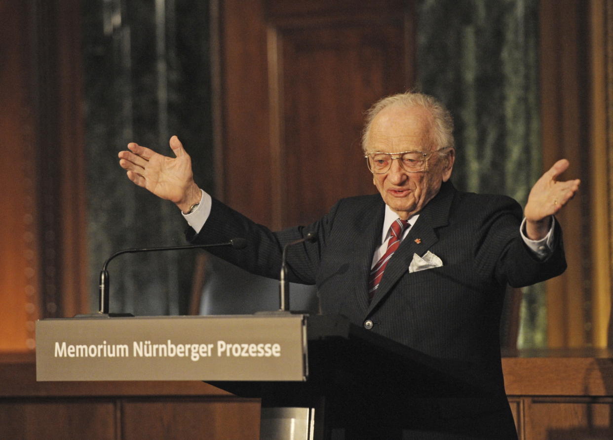 Benjamin Ferencz stands at a podium with his arms outstretched at the opening ceremony for the exhibition commemorating the Nuremberg war crimes trials.
