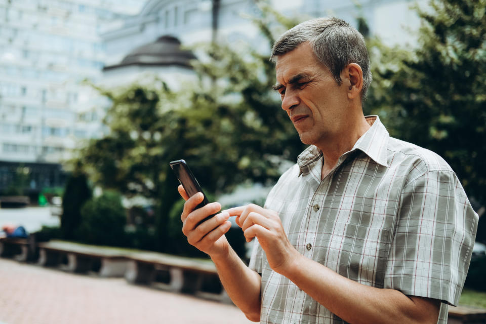 A man in a casual plaid shirt is standing outdoors, concentrating as he looks at his smartphone