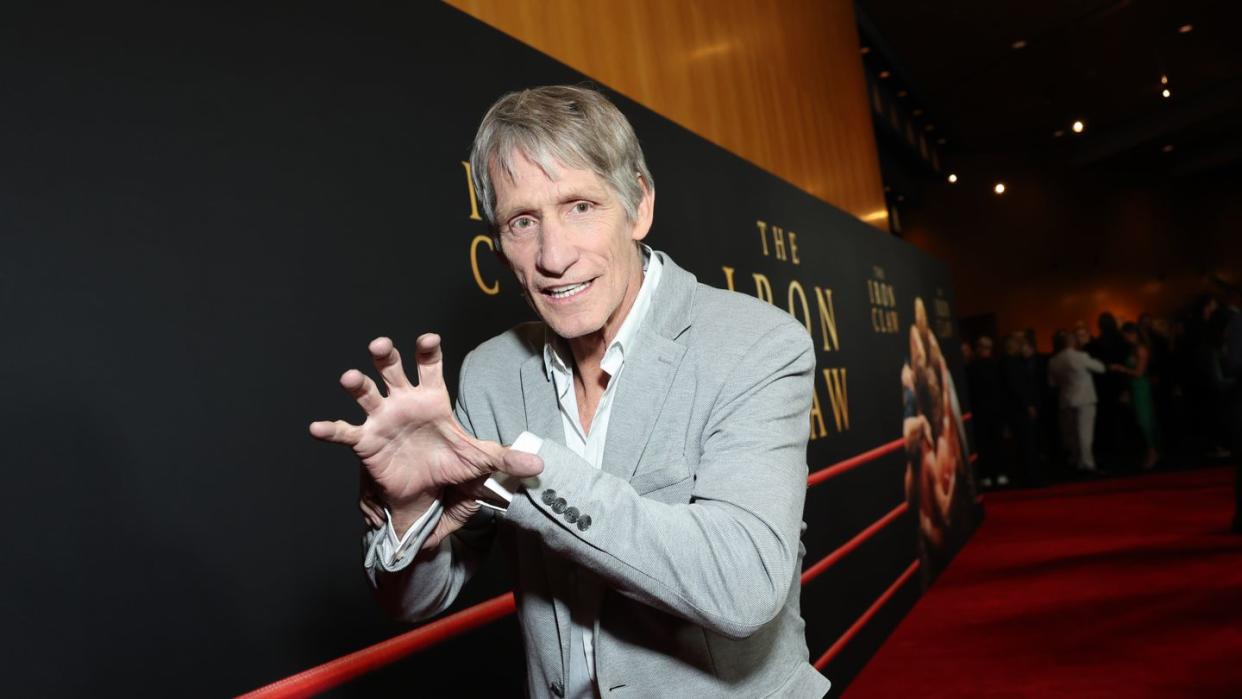 kevin von erich mimics the iron claw grapple for a photo while standing on a red carpet, he wears a gray suit jacket, white collared shirt and black pants, behind him is a large movie poster for the iron claw