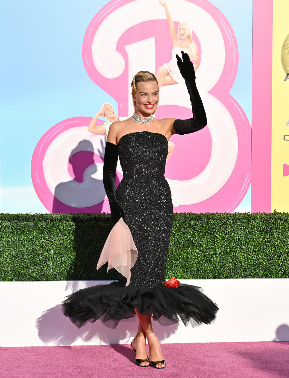 Margot Robbie in a black gown with ruffled detail, waving on the 'Barbie' premiere backdrop