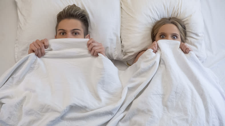Two people peeking out from under a bedsheet, expressions of surprise or concern on their faces