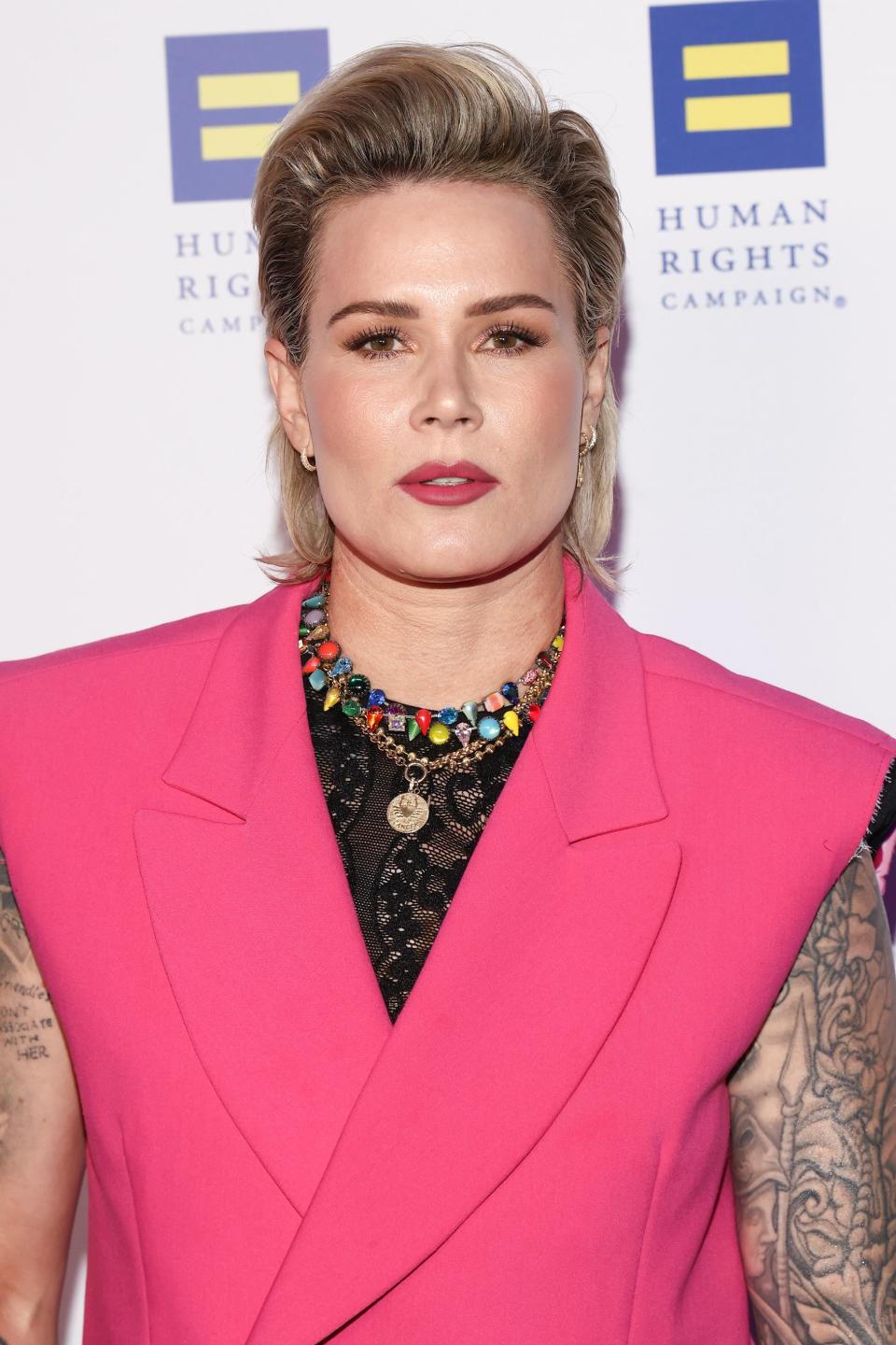 Ashlyn Harris in a pink blazer over a black top, adorned with a colorful necklace, with visible tattoos on the arms