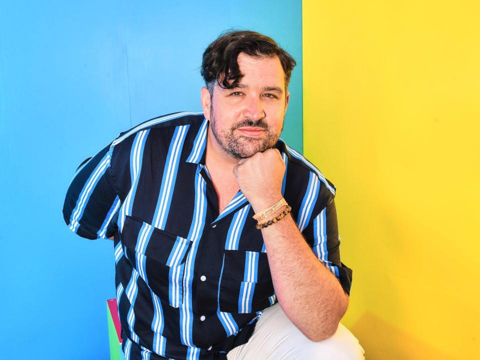 Brock Powell kneeling with his hand until his chin in a contemplative pose while wearing a bright blue and navy blue striped button-down shirt.