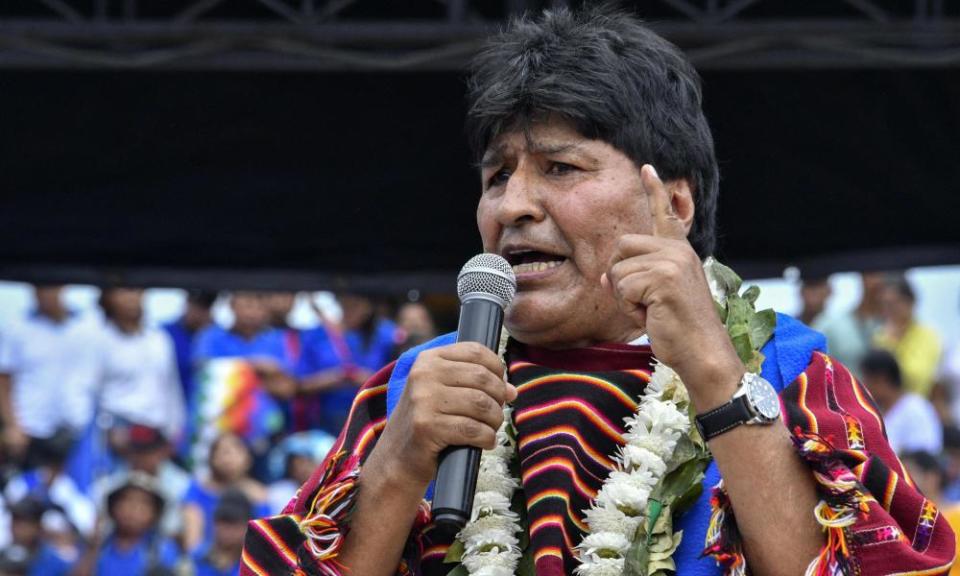 A middle-aged Bolivian man with thick black hair and wearing a red-and-orange ceremonial robe holds a microphone and points to the sky with his left hand as he speaks.