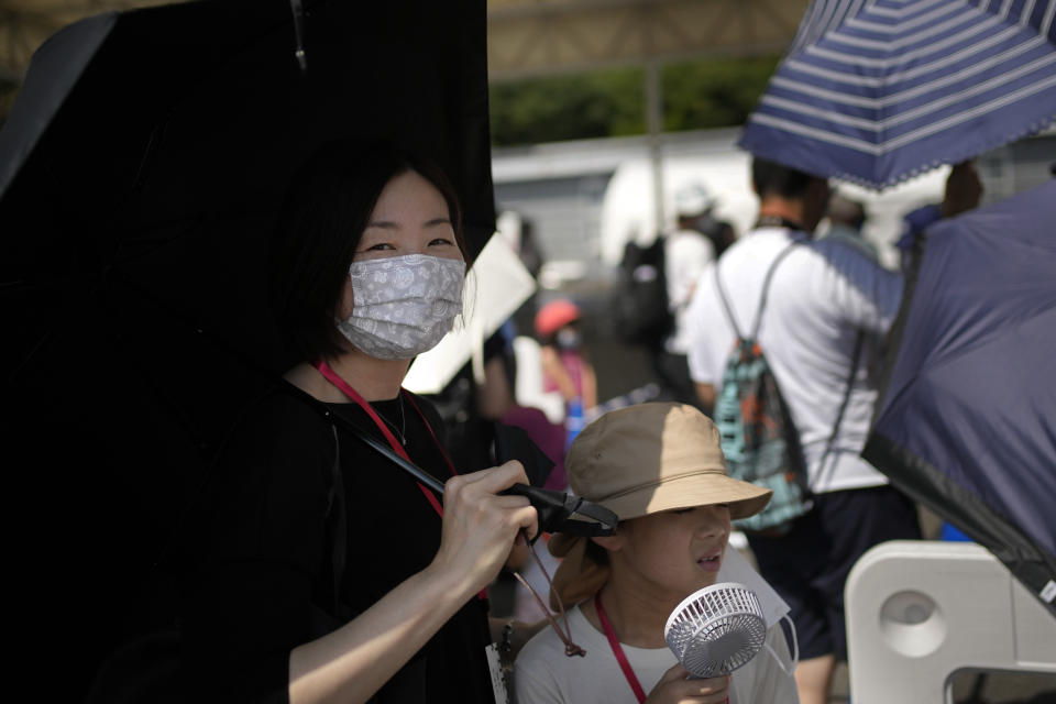 Visitors use umbrellas and fans to beat the heat outside the Fuji International Speedway, the finish for the women's cycling road race that is underway, at the 2020 Summer Olympics, Sunday, July 25, 2021, in Oyama, Japan. (AP Photo/Christophe Ena)