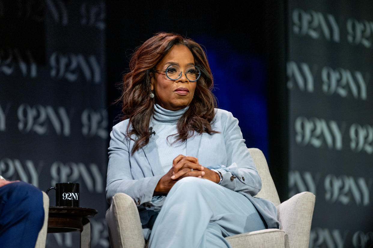 Oprah Winfrey seated with hands folded, wearing a pale blue suit and turtleneck.